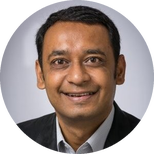 Vikas smiling in a dark blazer poses for a professional headshot against a light gray background.