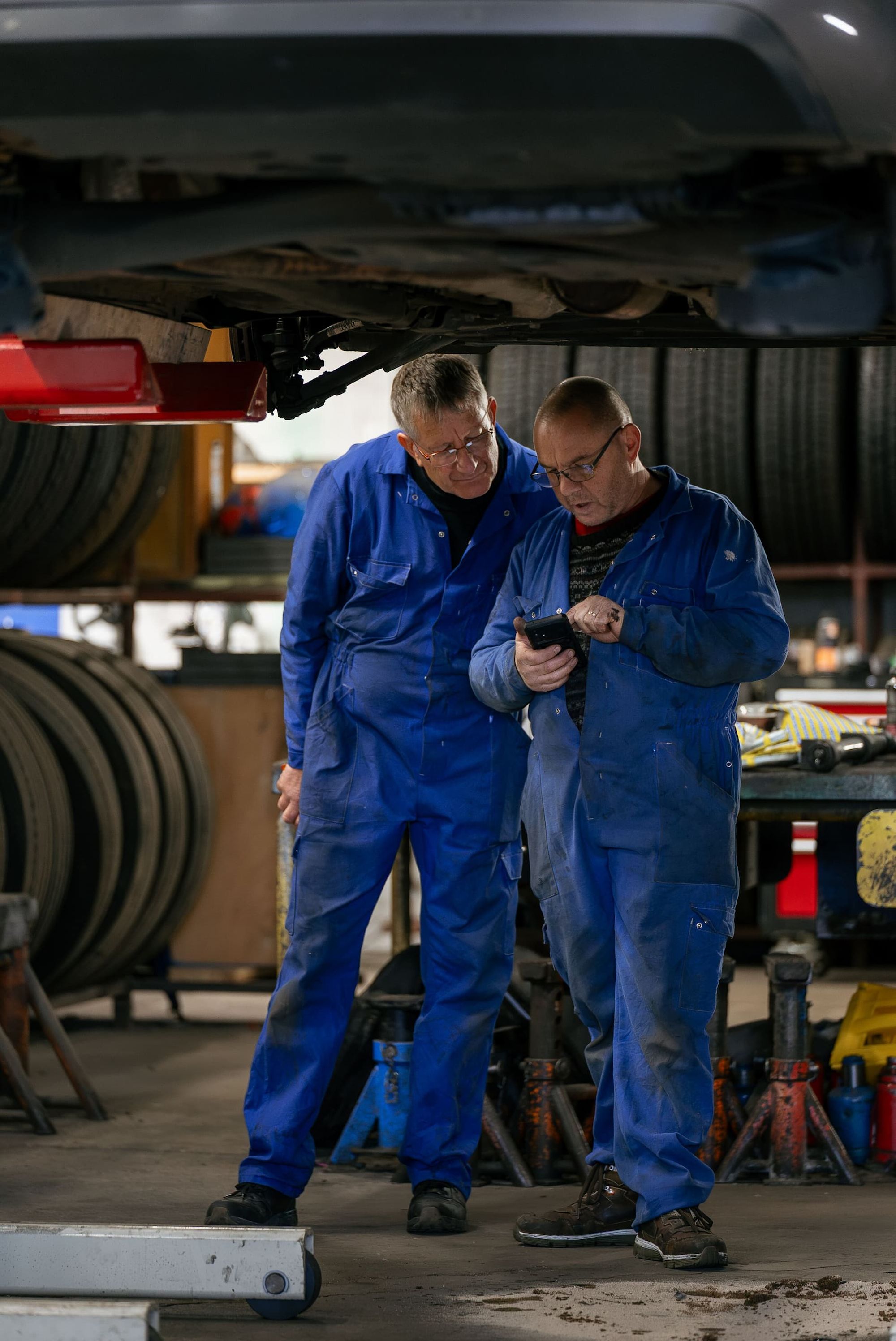 Two mechanics in blue coveralls review a smartphone under a lifted vehicle in an auto repair shop.