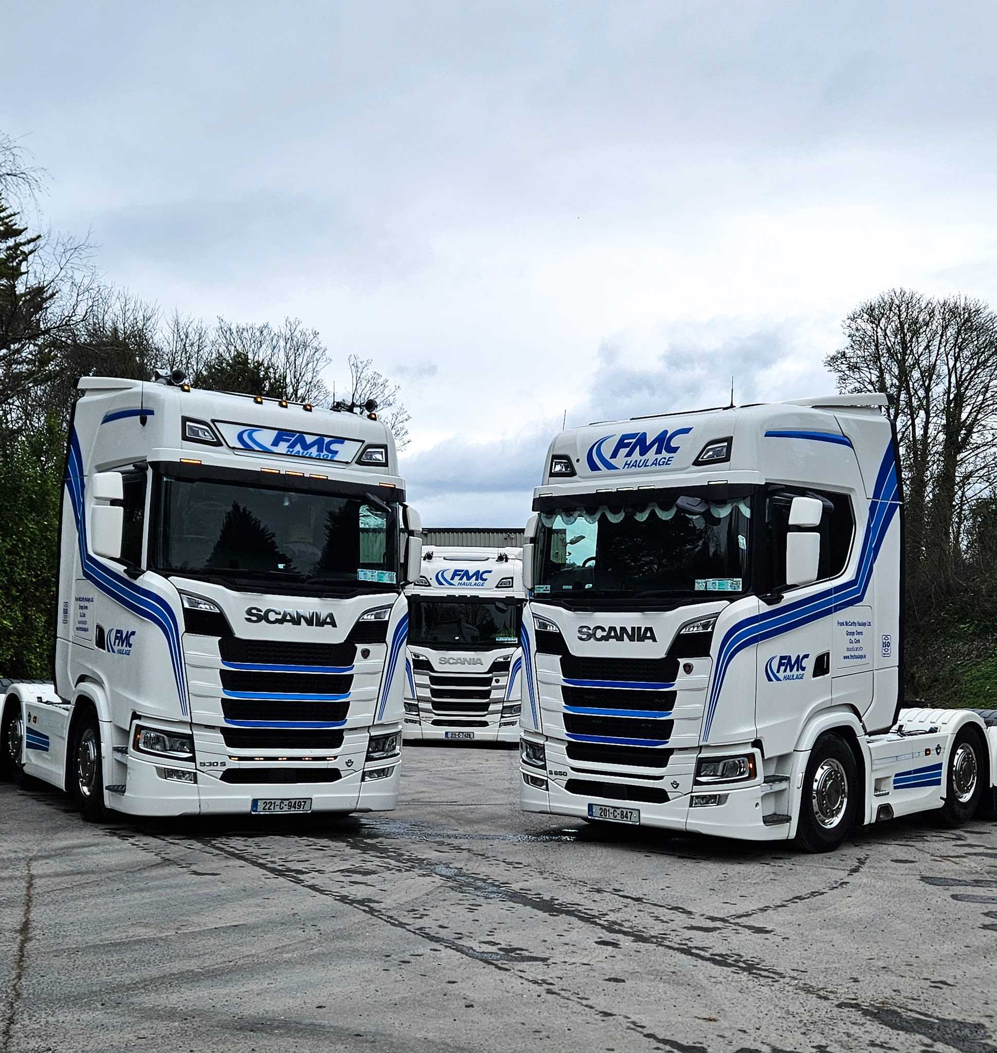 Three white Scania trucks with blue FMC Haulage livery parked in a yard under a cloudy sky.