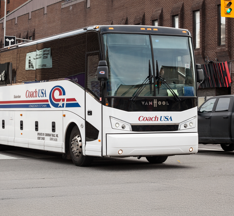 White Coach USA tour bus parked on a city street with brick buildings in the background and a traffic light visible.
