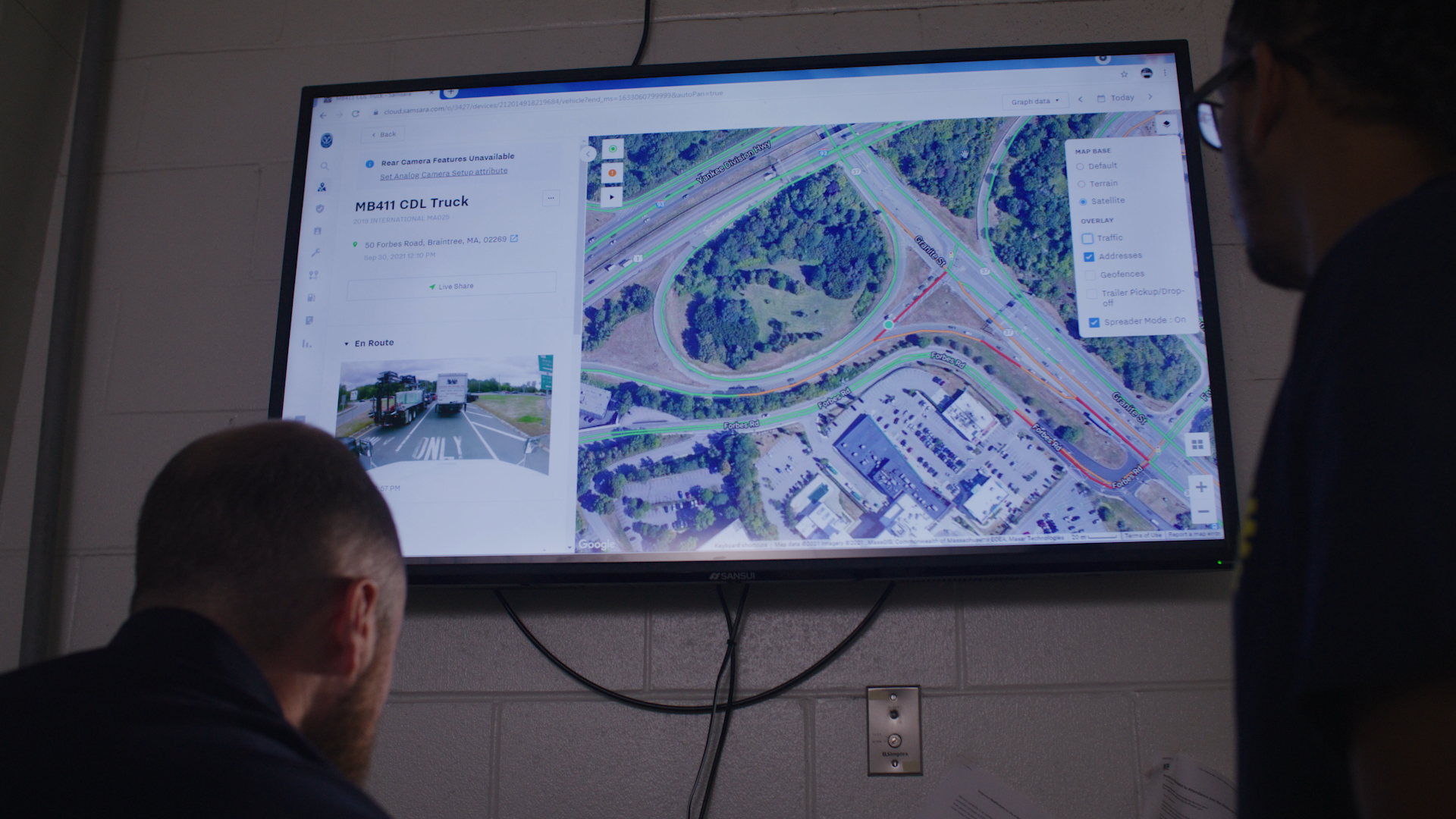 Two people view a monitor displaying a fleet tracking dashboard with a satellite map showing a CDL truck's route.