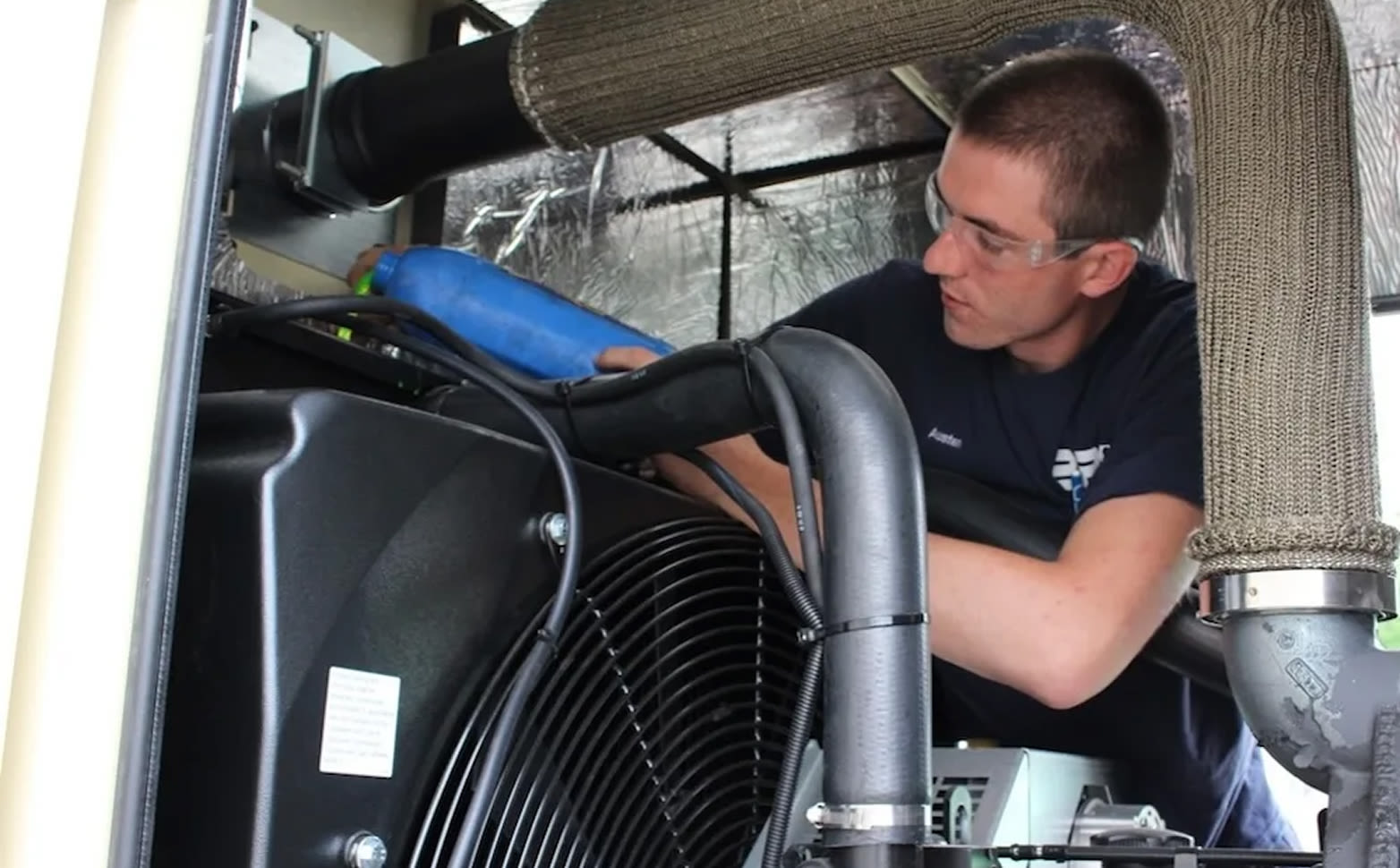 A technician holding a blue bottle inside a machinery compartment.