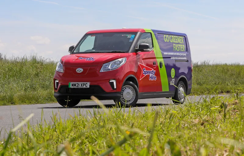 A red, purple, and green Speedy Hire electric van on a road, with "Go Greener Faster" text on the side.