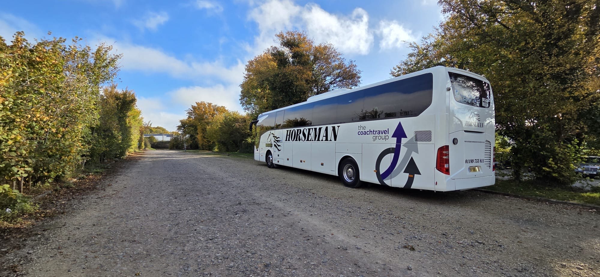 A white Horseman coach parked on a gravel road surrounded by autumn trees under a blue sky.