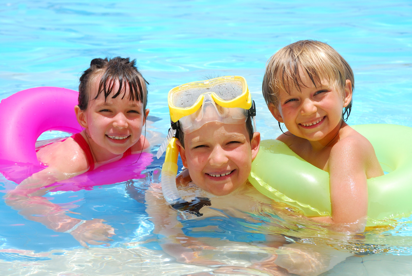 Three laughing children in the water with diving goggles and swimming tires.