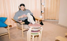 This image depicts a woman in her 20s, indicative of Generation Z, relaxing in an armchair while listening to her headphones and scrolling her phone.