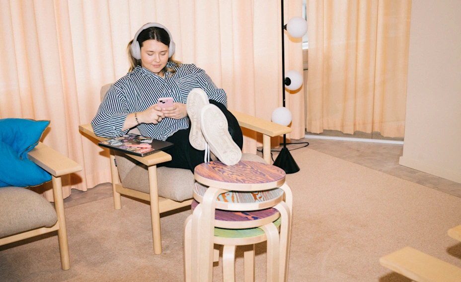 This image depicts a woman in her 20s, indicative of Generation Z, relaxing in an armchair while listening to her headphones and scrolling her phone.