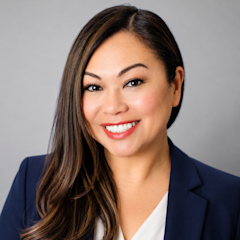 Headshot of Ann Yap with long brown hair smiling warmly and wearing a navy blazer with red lipstick, against a gray background.