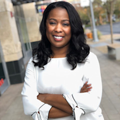 Headshot of Erica Rutherford with long black wavy hair, wearing a white blouse, standing outdoors with arms crossed.