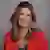 Headshot of Suzy Martinek, wearing a red blazer and necklace, posed against a gray brick wall background.