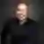 Aaron Gonzalez smiling in a black dress shirt, posed against a dark gray studio background.