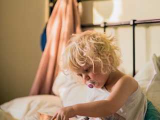 Kind mit Schnuller sitzt auf einem Bett und bedient ein Tablet