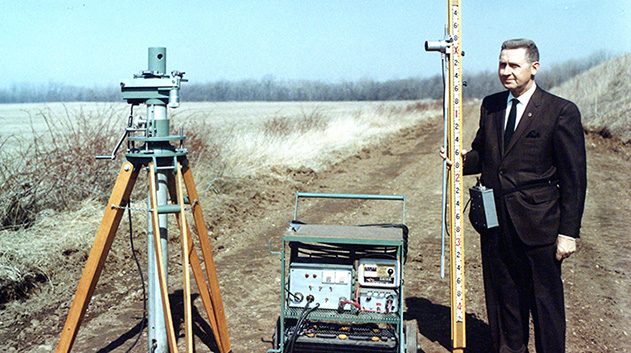 A historical photograph of a man in a suit and tie standing on a dirt road in a rural setting, holding a tall, calibrated surveying rod.
