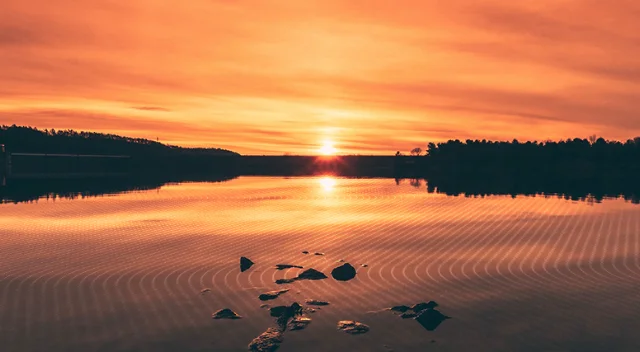 Golden sunset over massive dam, reflecting brightly on vast reservoir with concentric ripples. Dam silhouetted against sky.