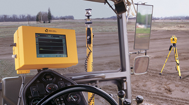 A view from inside the cab of a piece of heavy machinery, looking out at a dirt field. A large, yellow digital display screen is mounted to the right of the driver's position.
