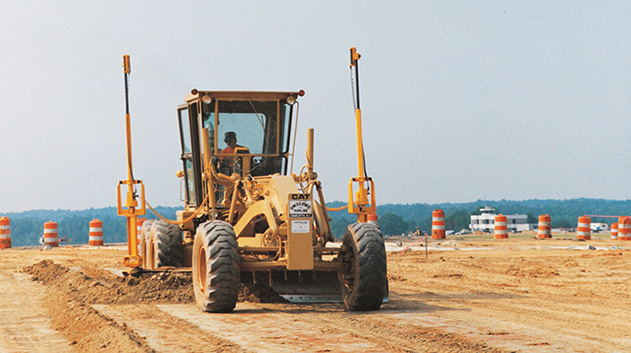 A medium shot of a large, yellow bulldozer or grader with a driver in the cab, moving dirt on a construction site.