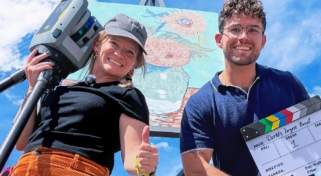 Two people pose at the World's Largest Easel in Goodland, Kansas, with a Trimble X9 laser scanner and a film clapperboard.