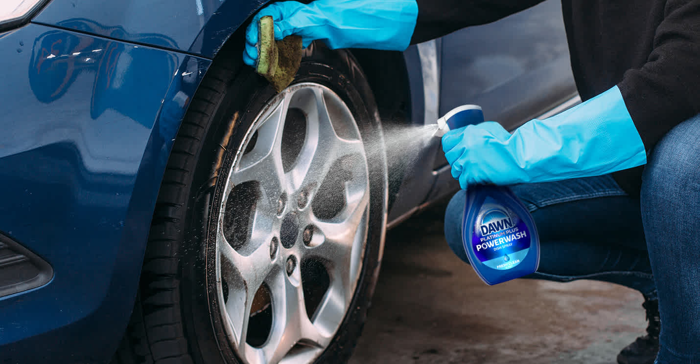 A person wearing blue rubber gloves spraying Dawn Powerwash dish spray onto the rim while wiping it with a green sponge