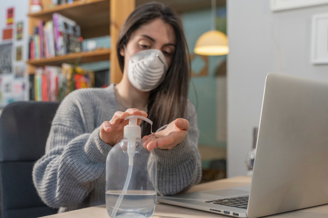 Woman at desk wearing mask and using hand sanitizer - Shutterstock