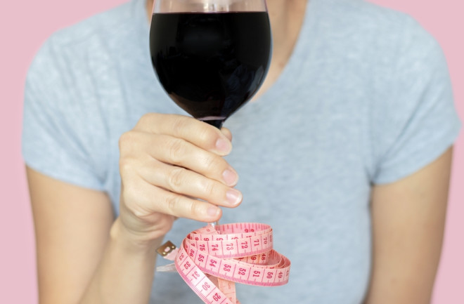 woman holding glass with red wine in hand tape measure attached by foot base. celebrating prohibited alcohol consumption. isolated girl pink background