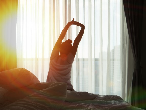 A woman waking up in the morning, stretching with the sun shining in through the window.  