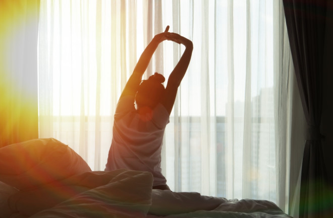 A woman waking up in the morning, stretching with the sun shining in through the window.  
