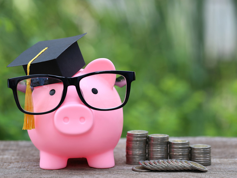 Pink piggy bank wearing a graduation mortar board and black glasses sitting next to 4 descending stacks of coins against a backdrop of trees.