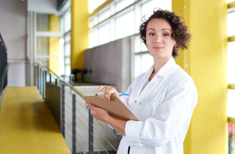 Young white female nurse with short brown curly hair wearing a white lab coat and holding a clipboard while standing in an open room with bright yellow walls and large windows.