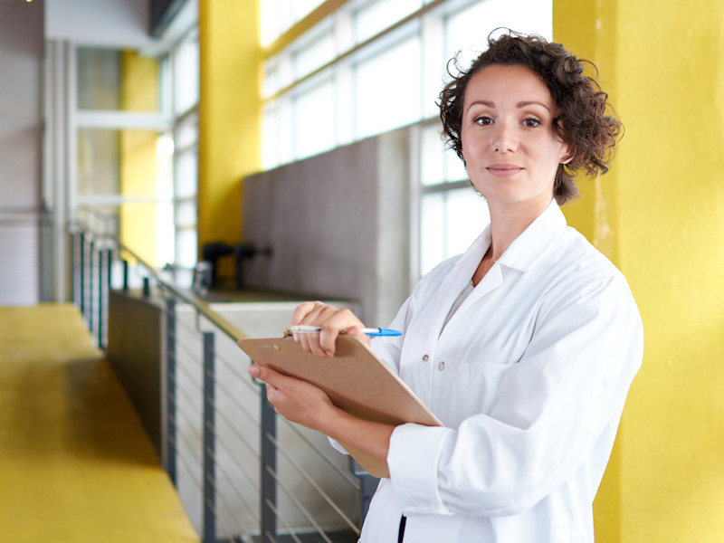 Young white female nurse with short brown curly hair wearing a white lab coat and holding a clipboard while standing in an open room with bright yellow walls and large windows.
