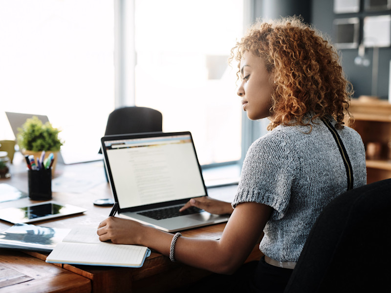 Young African-American woman sitting at a desk writing in a notebook while doing research on her laptop.