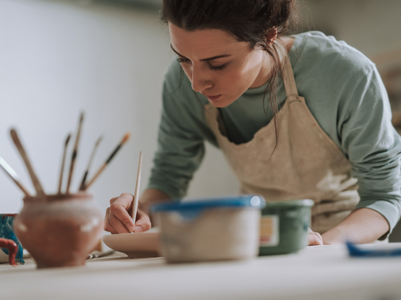 Young white woman in apron painting pottery in her workshop.