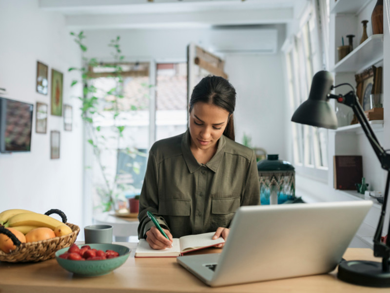 Young woman sitting at desk writing in notebook with laptop open in front of her.