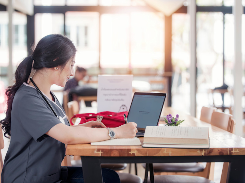 Female Asian-American nursing student wearing gray scrubs sits in a cafe doing her coursework on her laptop surrounded by books and notes.