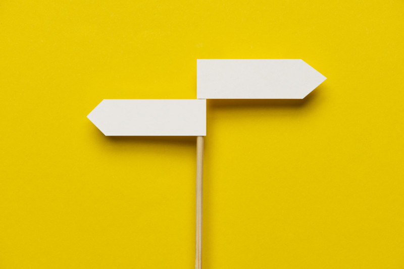 Skinny wooden pole with two white direction signs pointing in opposite directions against bright yellow backdrop.