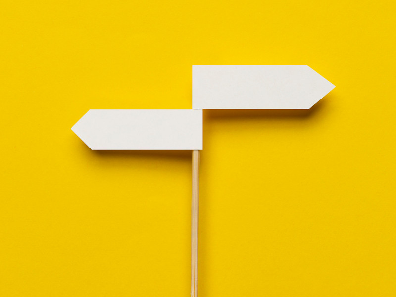 Skinny wooden pole with two white direction signs pointing in opposite directions against bright yellow backdrop.