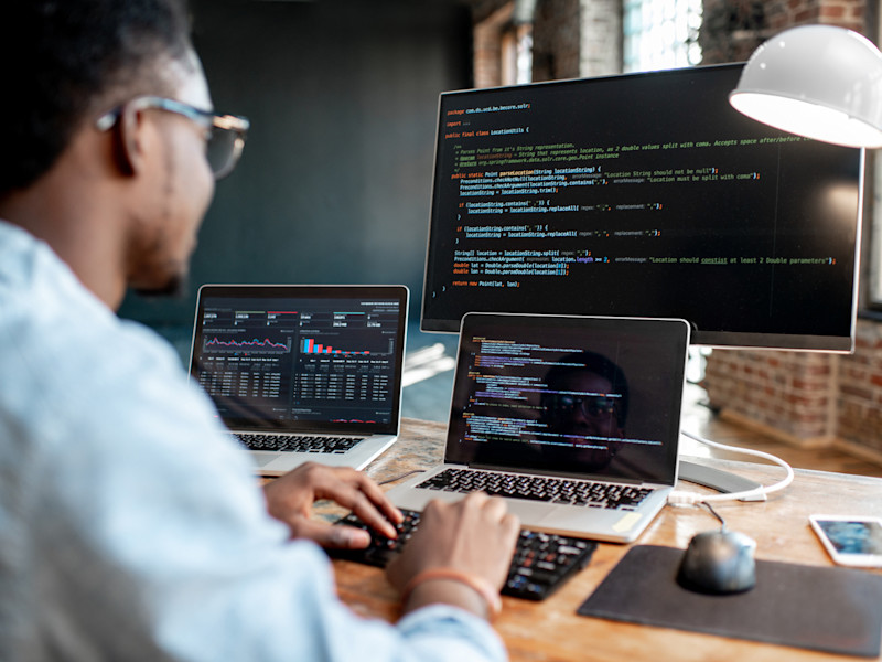 Profile of a young African-American programmer sitting at his desk with 3 monitors writing code.