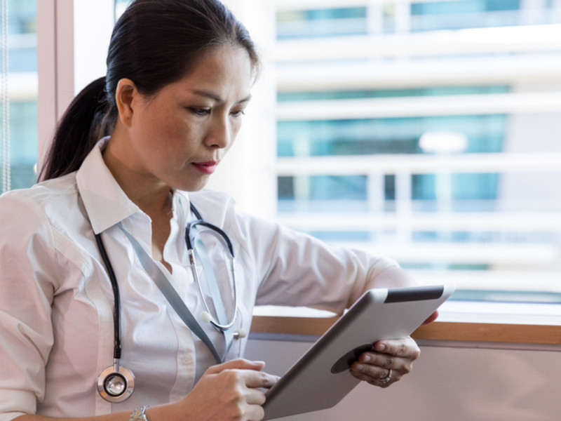 Asian-American female healthcare provider wearing a stethoscope and sitting in the window of a hospital reviewing her patients' charts on a tablet.