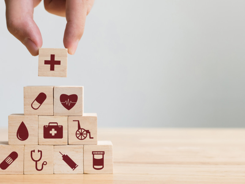 White hand putting a wooden block with a cross on top of a pyramid of wooden blocks with different medical-related icons on them.