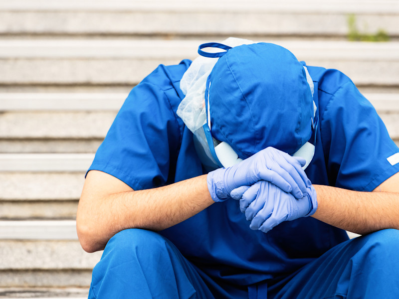 Stressed male healthcare workers in blue scrubs and cap with head on hands