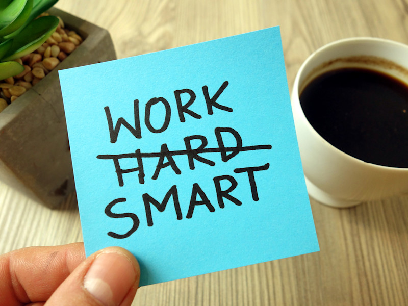 Hand holding a blue sticky note that says "Work Hard Smart" with the word "hard" struck through.