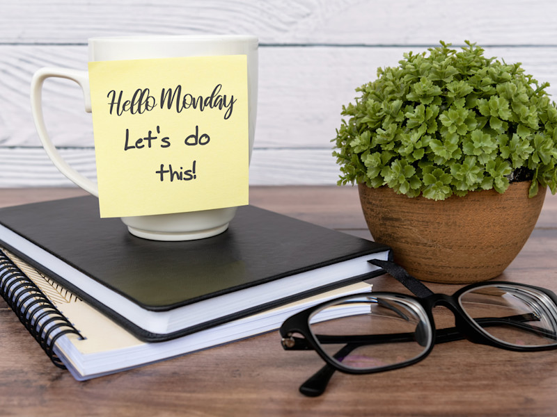 Coffee cup with a sticky note that says "Hello Monday let's do this" sits on top of two books and surrounded by a pair of black glasses and a decorative shrub, all on top of a brown desk.