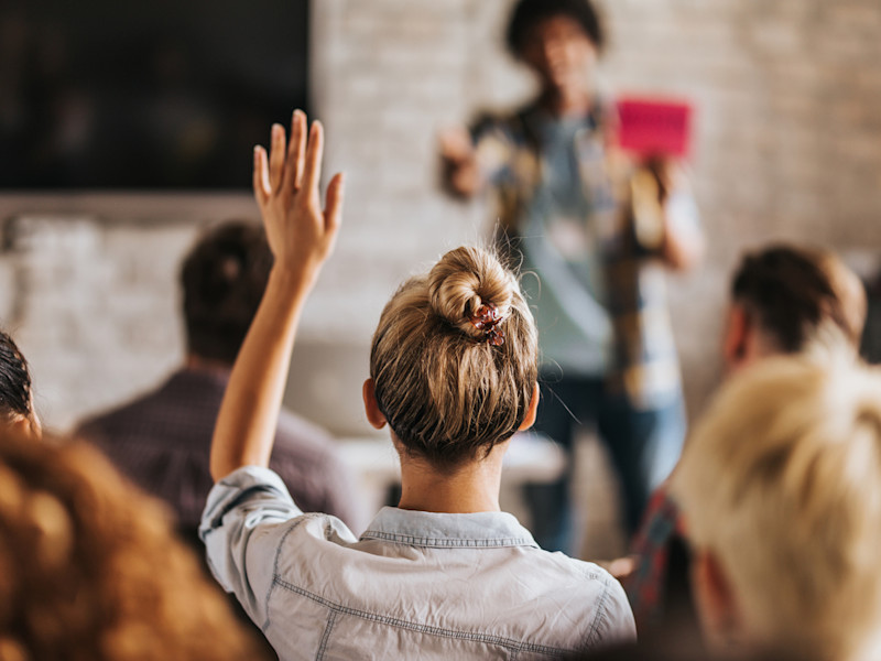 Looking at the back of a young blonde woman raising her hand in the middle of a crowded seminar.