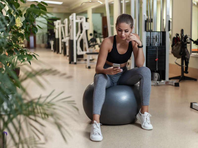Young female physical therapist in activewear sits on gray yoga ball in the middle of a gym while checking her smartphone.