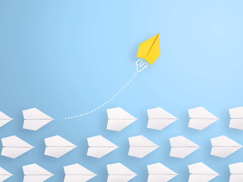 One yellow paper airplane breaking away from the pack of white paper airplanes flying against a baby blue background.