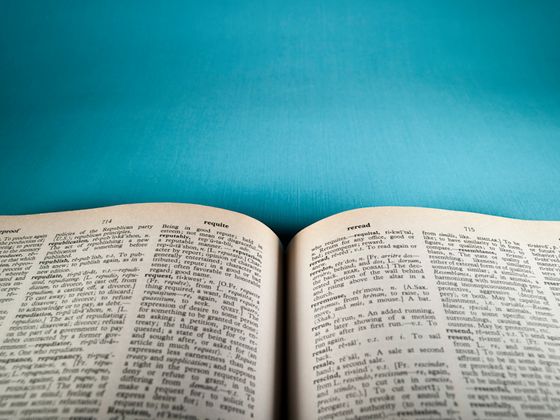 Slightly blurry view of a dictionary sitting open and showing two pages of definitions against a teal background.