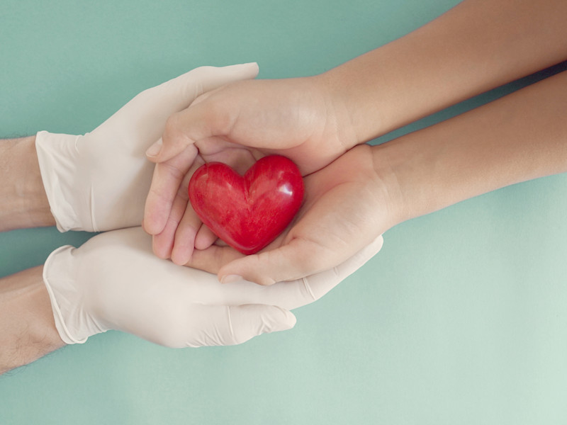 A pair of latex gloved hands holding bare hands that hold a red heart