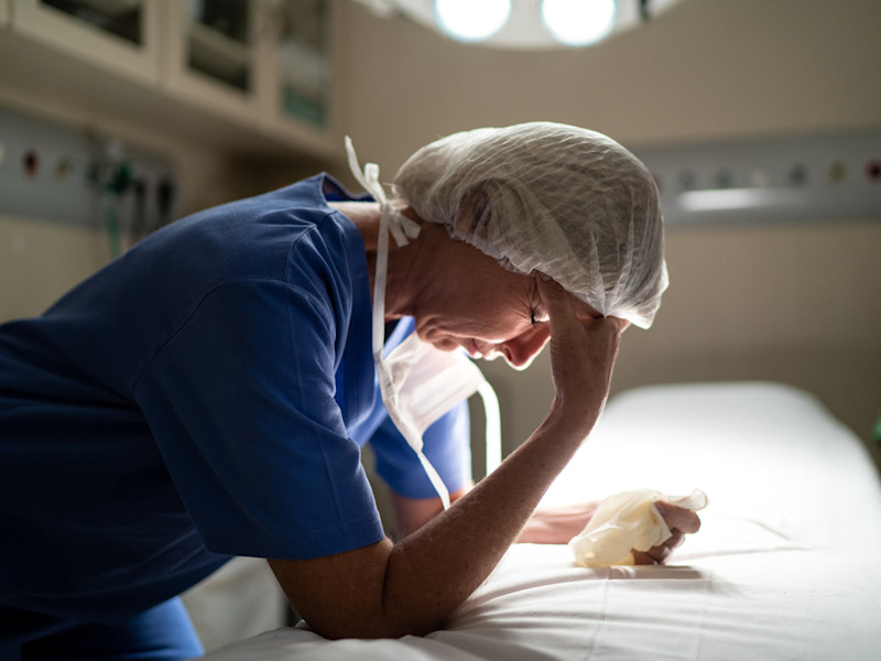 Stressed female nurse in PPE leans on bed and rubs forehead