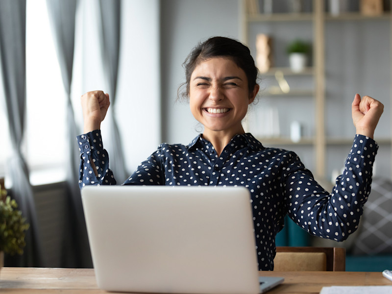 Young woman of color raising her arms in excitement while sitting in front of laptop.