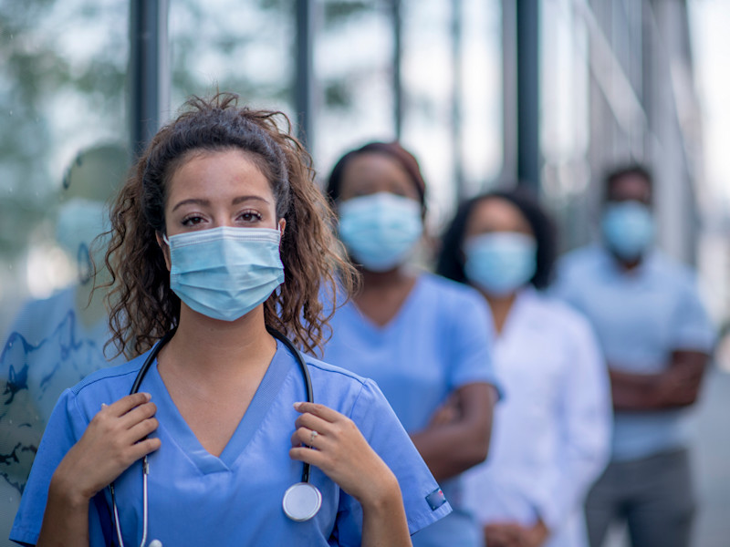 Masked female nurse practitioner standing outside facing camera with row of NPs standing behind her.