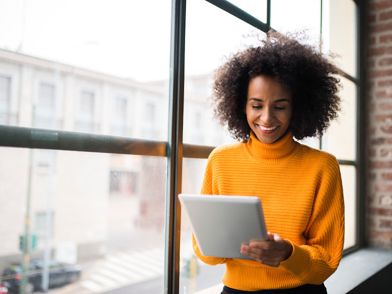 Female African-American real estate agent wearing an orange sweater and smiling while browsing on her tablet.